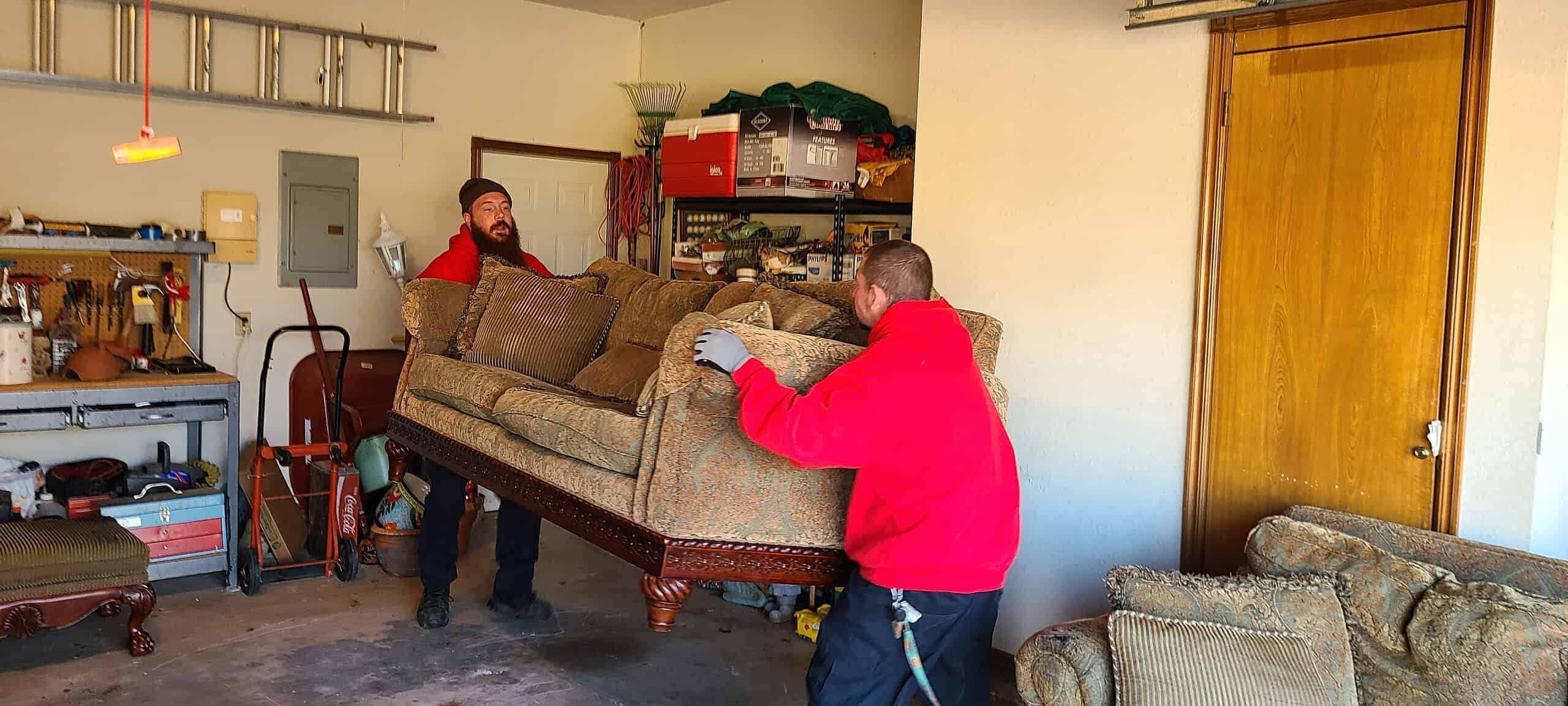 Two Junk Platoon crew members in red shirts carrying a couch out of a garage during a cleanout