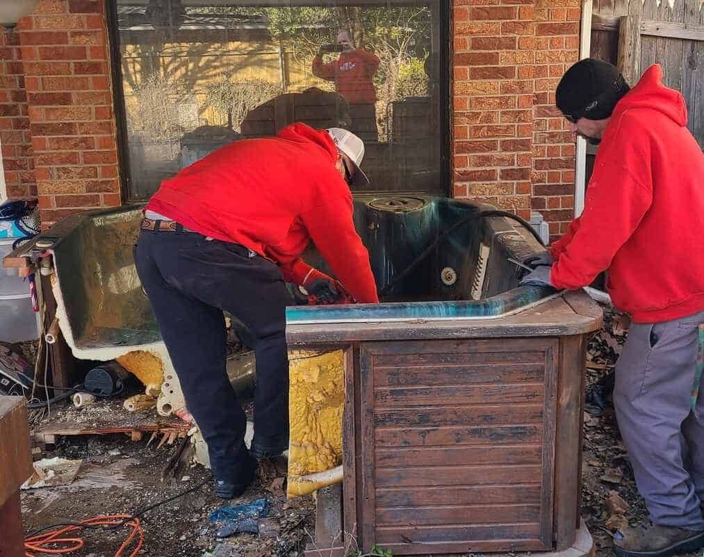Two Junk Platoon crew members in red hoodies cutting apart a hot tub for removal in Edmond, Oklahoma