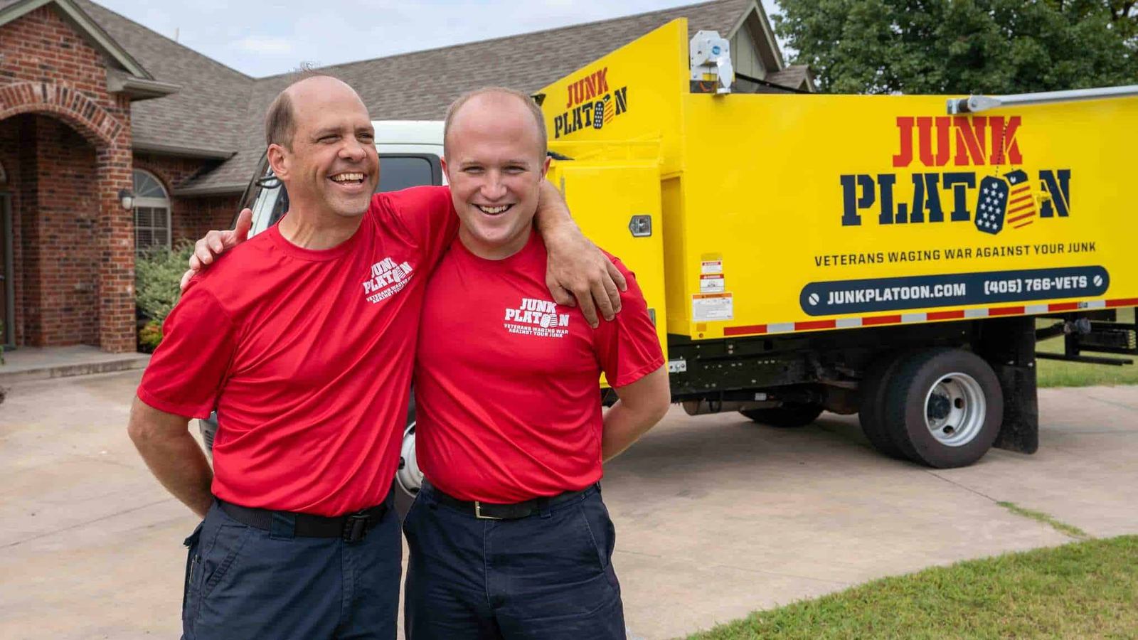 Two men in red Junk Platoon shirts standing arm in arm in front of a yellow Junk Platoon truck outside a brick home